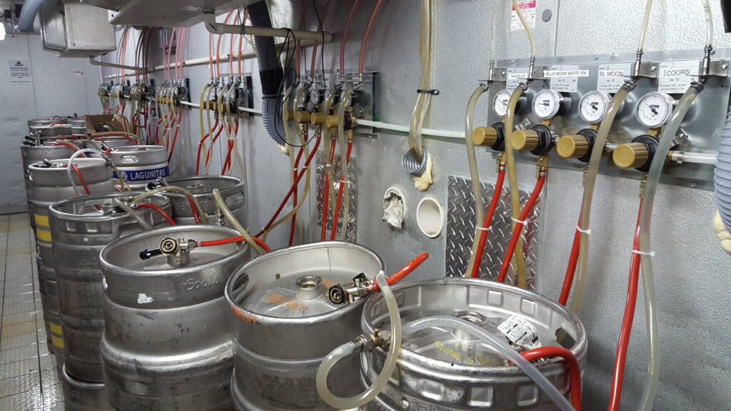 Several stainless steel kegs are connected to a system of tubes, gauges, and valves in a cooler room. The setup appears to be part of a draft beer distribution system.