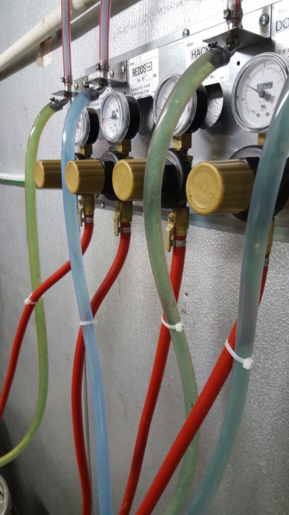 A close-up view of a wall-mounted panel with gauges and colored hoses, featuring brass fittings and clear tubes in red, green, and blue, likely part of an industrial or laboratory setup.