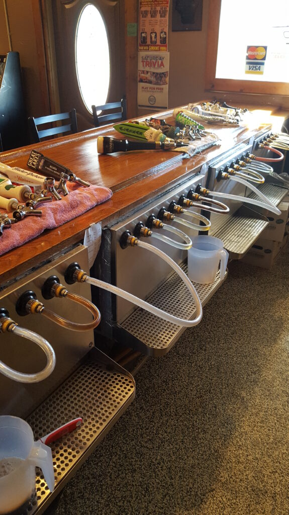 A bar setup showing multiple beer taps with hoses attached, plastic jugs underneath, and cleaning items on a wooden counter.